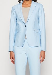 Blazer sur mesure bleu clair avec un bouton unique, deux poches avant, revers crantés et détail de logo subtil sur la poche. Texture de tissu lisse.