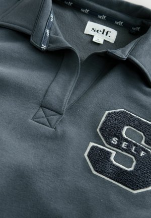 Gray polo shirt with a textured, embroidered "S" logo and "SELF" in white, featuring a classic collar and triangular stitching detail.