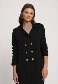 Czarny podwójnie zapięty płaszcz z złotymi guzikami, płaskim kołnierzem, długimi rękawami i bocznymi kieszeniami. Gładka tekstura, długość do kolan.