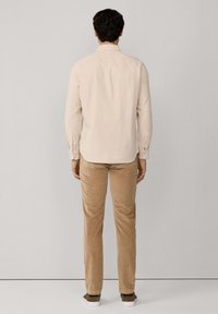 Chemise beige clair à manches longues avec un col boutonné, associée à un pantalon en velours côtelé beige et des baskets sombres, vue de dos.