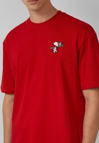 Rotes Baumwoll-T-Shirt mit kurzen Ärmeln, das ein gesticktes Snoopy-Design auf der linken Brust zeigt. Klassischer Rundhalsausschnitt und lässiger Schnitt.