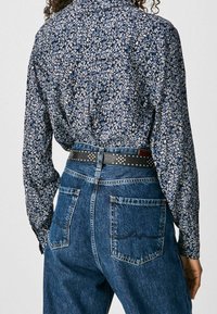 Kvinde iført en langærmet skjorte med blå blomster, som er stoppet ned i højtaljede blå jeans med et sort bælte med sølvnitter.