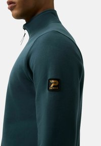 Polarowy pulower w ciemnym odcieniu tealu z wysokim kołnierzem, z czarną i złotą naszywką logo na rękawie. Gładka faktura, dopasowany krój.