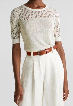 Osoba ubrana w białą dzianinową bluzkę z krótkim rękawem z geometrycznym wzorem oraz białe spodnie z wysokim stanem i brązowym paskiem.