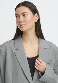 Grå blazer lavet af glat stof, med en enkelt brystlomme, strukturerede revers og afslappet pasform med let oversize ærmer.