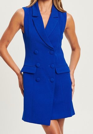 Femme portant une robe blazer bleu roi sans manches, à double boutonnage, avec des revers crantés et des poches à rabat, debout les mains près des hanches.