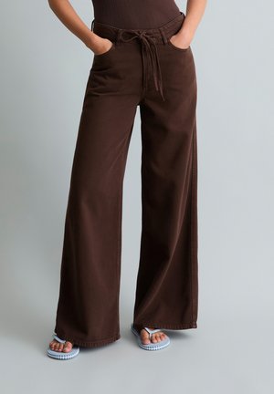 Personne portant un pantalon large marron foncé avec une ceinture à cordon et des tongs bleues, debout les mains dans les poches sur un fond clair.