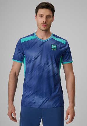 Mężczyzna w niebiesko-turkusowej koszulce sportowej z krótkimi rękawami i geometrycznymi wzorami, zwrócony przodem na jednolitym szarym tle.