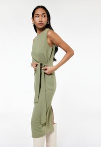 Vestido sin mangas de color verde oliva con cintura atada, abertura lateral y textura suave. Combinado con botas blancas hasta la rodilla, sobre un fondo claro.