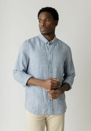 Mand med kort krøllet hår iført en lyseblå linned skjorte med knapper og beige bukser, stående med hænderne foldet, ser til siden.