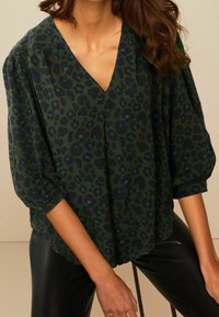 Blouse à imprimé léopard vert avec un col en V, une coupe ample et des manches bouffantes, réalisée en tissu léger à la texture lisse.