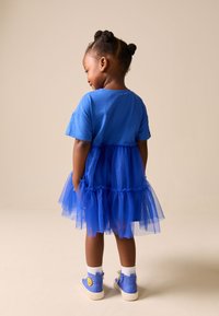 Jeune fille aux cheveux tressés portant une robe en tulle bleue et des baskets bleues avec un motif soleil, debout de profil sur un fond beige.
