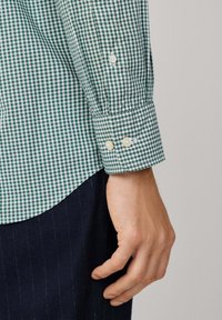 Antebrazo y mano de una persona que lleva una camisa de cuadros verde y blanca con puños abotonados, combinada con pantalones oscuros a rayas.