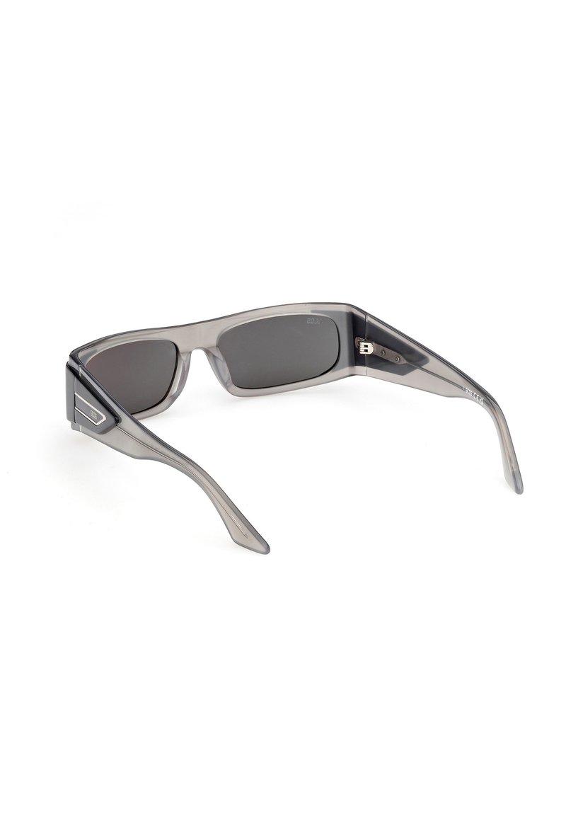 Rechthoekige zonnebril met donkere glazen, transparante grijze plastic monturen en brede pootjes, licht schuin geplaatst op een wit oppervlak.