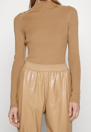 Femme portant un pull côtelé beige à manches longues avec des boutons aux épaules et un pantalon en cuir beige taille haute avec une ceinture élastique.