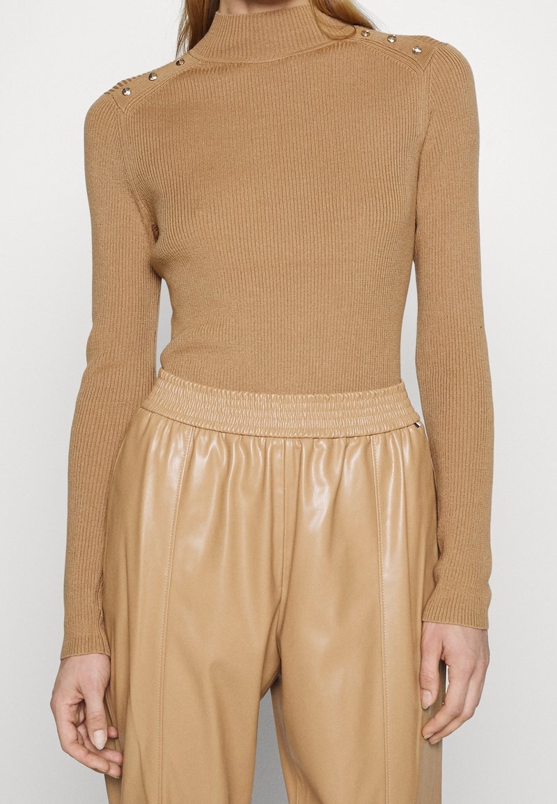 Femme portant un pull côtelé beige à manches longues avec des boutons aux épaules et un pantalon en cuir beige taille haute avec une ceinture élastique.