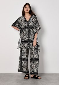 Schwarz-weiß gemusterter Kaftan mit weiten Ärmeln und lockerer Passform, kombiniert mit passenden weiten Hosen und schwarzen Sandalen.