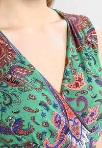 Färggklad grön klänning med djup V-ringning, intrikata paisley- och blommönster i rött, blått och vitt, med mjuk tygkänsla.