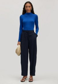 Blaues Langarmoberteil, tailliertes Design. Hochgeschnittene marineblaue Hose mit lockerer Passform. Hält eine beigefarbene Clutch; trägt schwarze Sandalen.