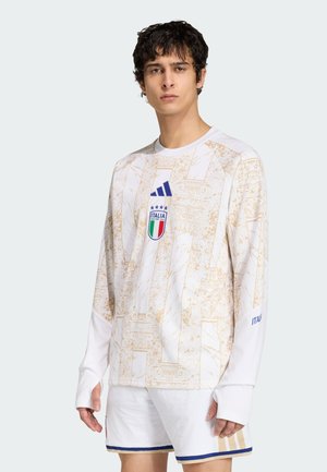 Joven con camiseta blanca de fútbol de Italia con patrones arquitectónicos dorados y logo, combinado con pantalones cortos blancos con ribete azul y dorado.