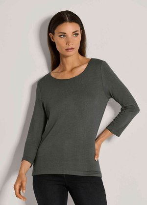 Frau mit langen dunklen Haaren, die ein dunkelgraues Dreiviertelarmshirt und schwarze Hosen trägt, steht mit einer Hand an der Hüfte.