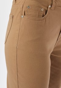 Zbliżenie na brązowe spodnie, ukazujące przednie i kieszeń na monety z metalowymi nitami oraz szczegółowym przeszyciem na materiale.