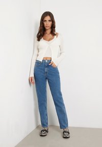 Cardigan scurt cu mâneci lungi, alb cu dungi, peste un top lejer, purtat cu blugi albaștri cu talie înaltă și încălțări slip-on cu model.