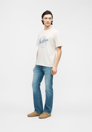 Young man wearing white graphic t-shirt, blue jeans, and tan sneakers, standing with hands relaxed, looking to the side on plain background.