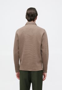Polo de manga longa castanho texturizado com colarinho. O design apresenta uma leve textura em ribas e mangas caídas, combinado com calças verdes.