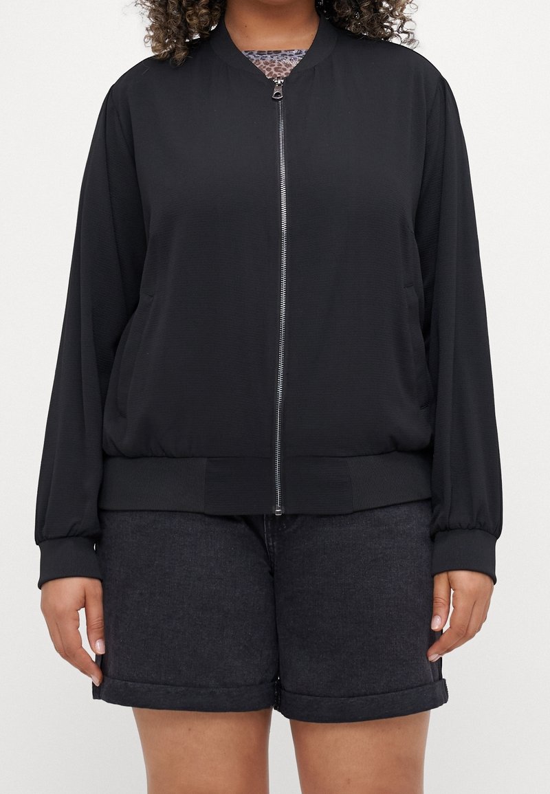 Femme portant une veste bomber noire zippée sur un top imprimé léopard et un short foncé avec ourlet roulé, debout, les bras détendus le long du corps.
