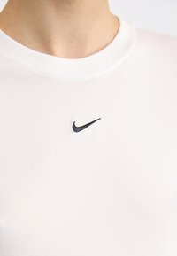 Bílý bavlněný tričko s kulatým výstřihem a malým černým logem Nike na hrudi, hladká textura bez vzorů nebo dalších detailů.