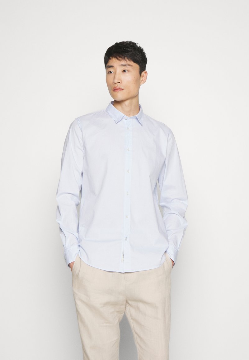 Chemise bleu clair à boutons avec col, manches longues et texture subtile, associée à un pantalon beige clair.