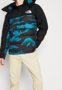 Schwarze und teal-blaue Steppjacke mit horizontalen Steppabschnitten, einem halben Reißverschluss auf der Vorderseite und einem Logo auf der Brust. Weicher Stoff und Kapuze.