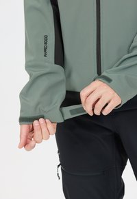 Softshell-jakke i dæmpet grøn med sorte accenter, med justerbare manchetter, en frontlynlås og logodetalje på ærmet.