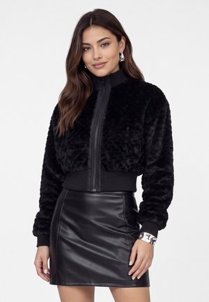 Femme aux longs cheveux bruns portant des boucles d'oreilles créoles argentées, une veste zippée noire texturée et une jupe en cuir noire, souriant légèrement.