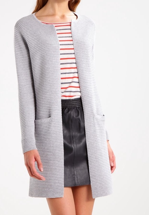 Vrouw in een lichtgrijze ribgebreide lange cardigan over een gestreept top en een zwarte leren rok tegen een witte achtergrond.