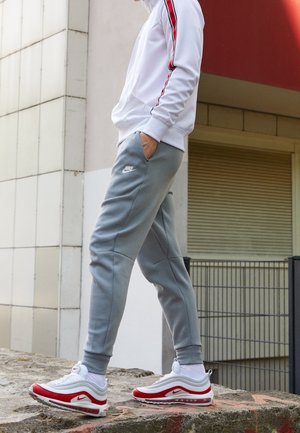 Person iført grå Nike joggingbukser, hvide og røde Nike sneakers, og en hvid jakke med rød stribe, står udendørs på en betonkant.