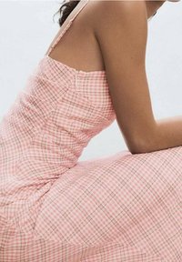 Růžové kostkované šaty s tenkými ramínky, které mají řasený design a jemný kostkovaný vzor. Látka vypadá lehce a má hladkou texturu.