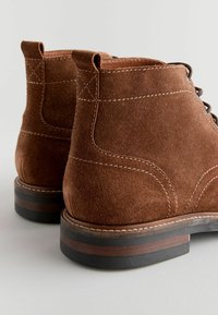 Braune Wildleder-Stiefeletten mit abgerundeter Spitze, kontrastierendem weißen Nähten, Lederakzenten an der Ferse und Zugschlaufen auf der Rückseite.