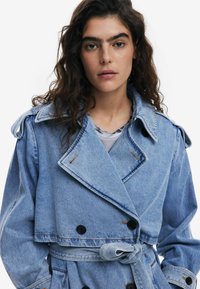 Denim-Trenchcoat mit cropped Design, hellblauer Farbe, großem Kragen, doppelter Knopfleiste und passendem Gürtel in der Taille.