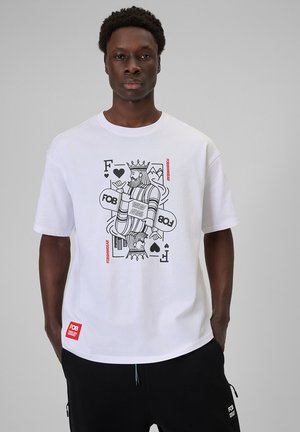 Młody mężczyzna w białym T-shircie z czarno-czerwonym nadrukiem króla z kart do gry oraz czarnych spodniach z kieszeniami.