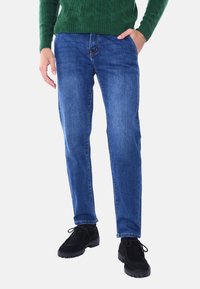 Jeans in denim blu con taglio dritto, caratterizzati da cinque tasche e dettagli di cucitura sottili, abbinati a scarpe in suede nere.