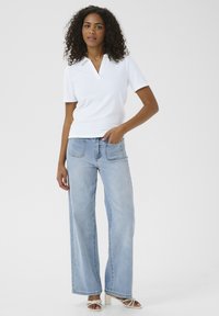 Weißes, geripptes Polo-Shirt mit kurzen Ärmeln, kombiniert mit hellblauen weiter geschnittenen Jeans. Das Model steht mit einer Hand in der Jeanstasche und präsentiert ein lässiges Outfit.