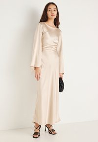 Ilga, smėlio spalvos satino suknelė su prigludusiu liemeniu, platėjusiomis rankovėmis ir lygiu audiniu. Juoda delninė rankinė ir juodi basutės su dirželiais papildo žvilgsnį.