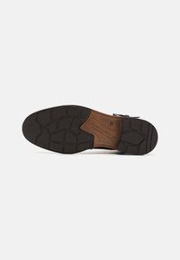 Suela de zapato de cuero marrón con goma negra texturizada, que presenta un dibujo en la banda de rodadura y un detalle de marca en relieve en la sección color marrón claro.