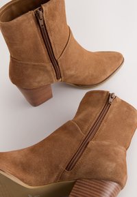 Enkellaarsjes van lichtbruin suède met een spits toelopende neus en een rits aan de zijkant. Stevige, gestructureerde houten hak. Gladde voering aan de binnenkant, minimale stikdetails.