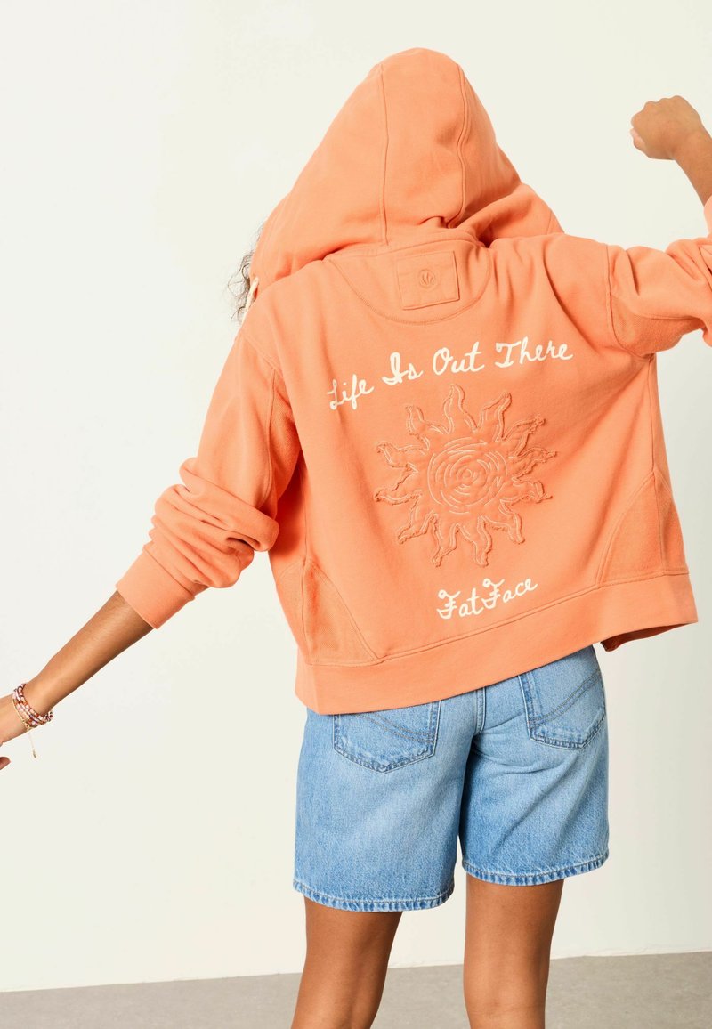 Személy narancssárga kapucnis pulóverben, nap motívummal és "Life Is Out There Fat Face" felirattal a hátulján, kék farmer rövidnadrággal párosítva.