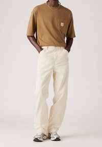 T-shirt marrone a maniche corte con tasca sul petto abbinato a pantaloni beige a vestibilità rilassata e sneakers grigie, dotate di lacci e dettagli texturizzati.