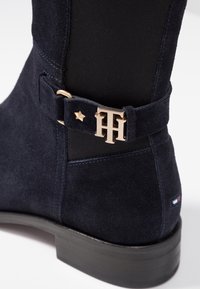 Stivale alla caviglia in suede blu navy con pannello elastico nero, fibbia con logo "TH" dorato, piccolo dettaglio a stella dorato e suola nera con una piccola etichetta rossa, bianca e blu.