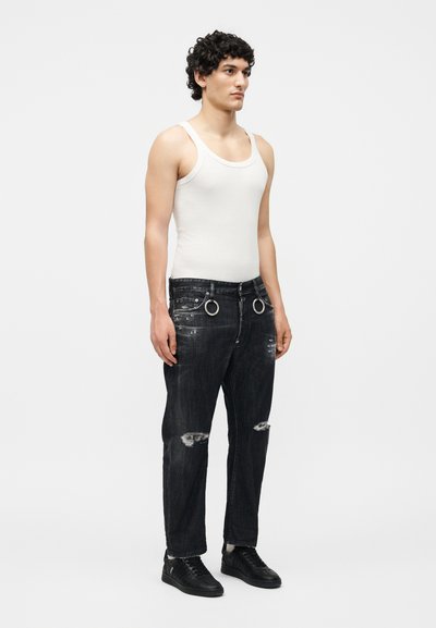 Άνδρας μοντέλο στέκεται φορώντας λευκό αμάνικο μπλουζάκι, μαύρα τζιν με σχισίματα και μεταλλικά δαχτυλίδια, και μαύρα αθλητικά παπούτσια σε απλό ανοιχτό φόντο.